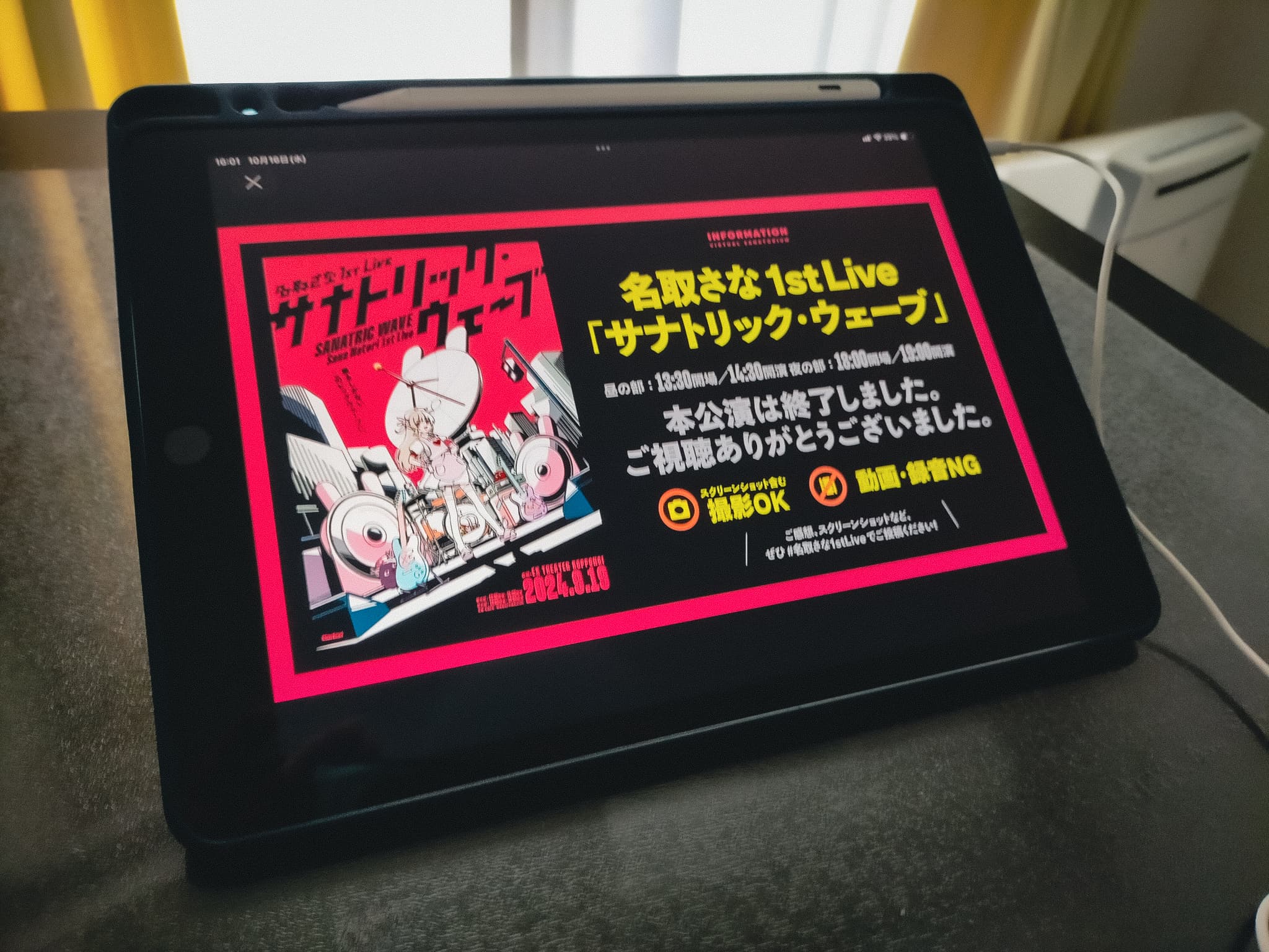 「名取さな1stLive「サナトリック・ウェーブ」」というタイトルがキービジュアルとともに画面に映っているiPadが机の上に置かれており、イヤフォンのコードが手前に伸びている。